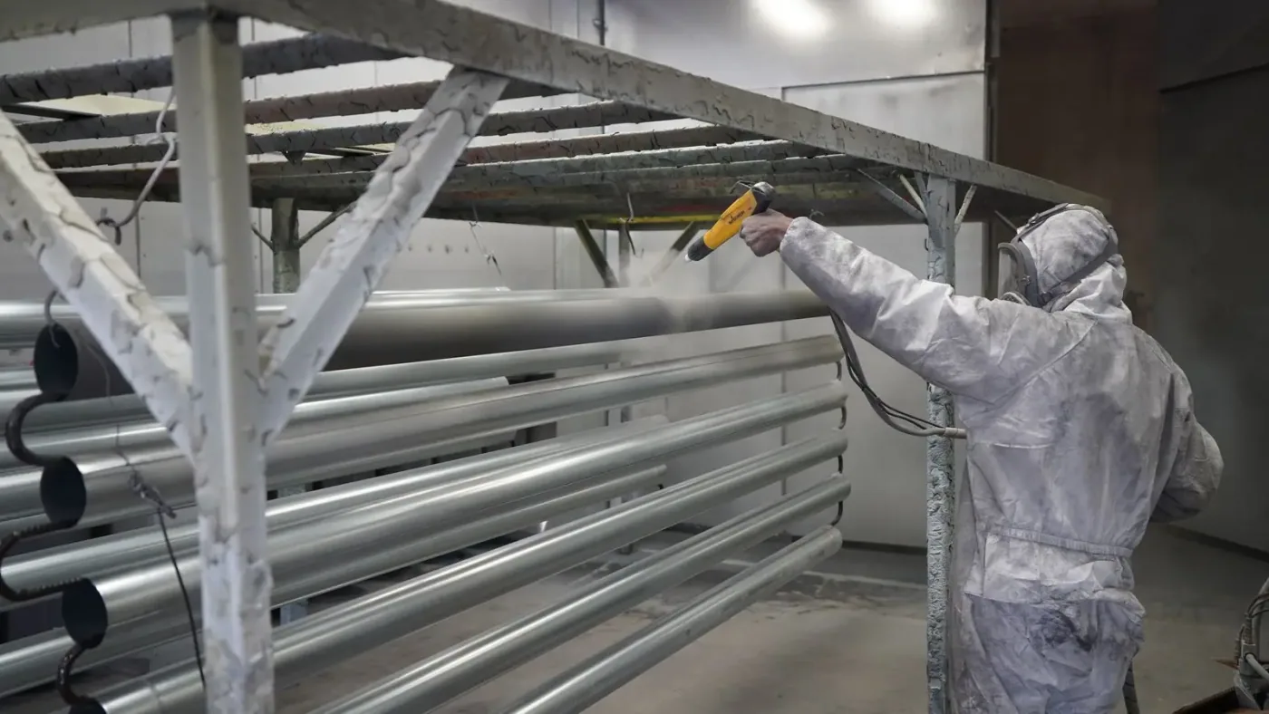 Operator in a protective suit spraying powder onto long aluminum tubes on a finishing rack