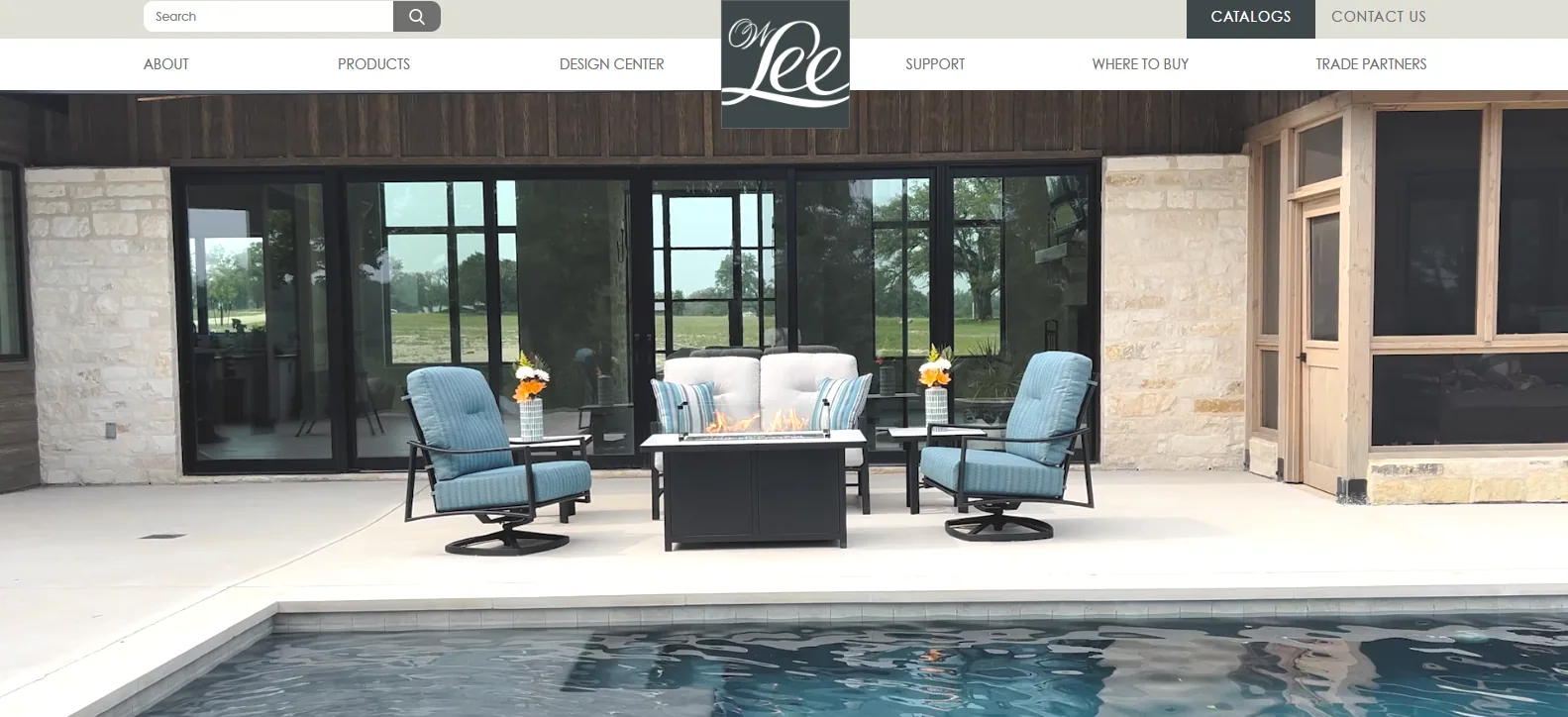 O.W. Lee homepage hero showing deep blue cushioned swivel lounge chairs and a sofa around a fire table on a concrete pool terrace, with its Design Center and Trade Partners navigation