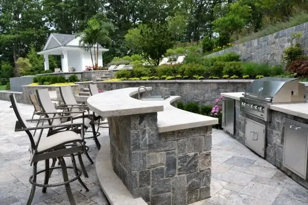 Curved stone outdoor bar counter with round bar stools facing a built-in stainless grill and outdoor kitchen cabinets on a paver patio